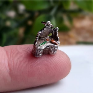 May include: A miniature silver vanity charm with an open mirror, drawers, and decorative detailing. The charm features small painted accents of green and red. The charm is approximately 0.75 inches in height.