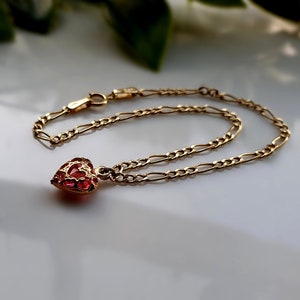 Può includere: Un bracciale a catena color oro con un ciondolo a forma di cuore contenente una pietra rossa, traslucida e sfaccettata, incastonata in una gabbia di filigrana dorata. Il bracciale presenta una chiusura a molla e maglie ovali allungate.