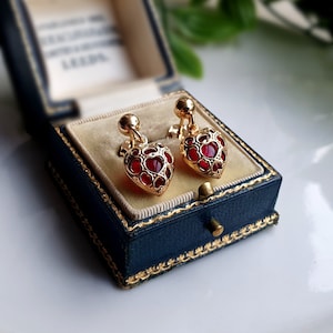 May include: Gold-coloured heart-shaped earrings with red detailing, presented in a vintage blue jewellery box. The earrings have a filigree design. The box has a light beige interior.