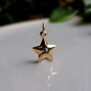 May include: A gold-toned star-shaped pendant featuring a small, circular blue gemstone set in the centre. The pendant includes a small circular jump ring at the top for attachment, with a smooth, polished metallic finish reflecting light.