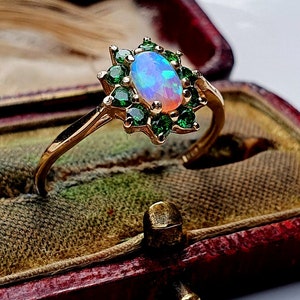 May include: A gold ring featuring a central oval-shaped iridescent opal surrounded by a halo of small, round, vibrant green gemstones. The setting is floral-inspired, with the green stones arranged in a petal-like pattern around the opal.