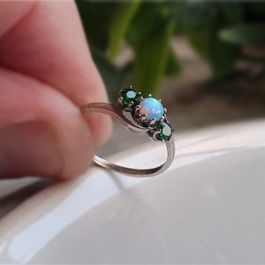 May include: A silver ring featuring an opal gemstone and two emeralds. The opal is the central focus, with a blue and white hue. The ring has a flowing design. The emeralds are a deep green colour.