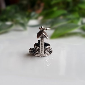 May include: Silver charm shaped like a well with a roof. The well is detailed with brickwork and a small bucket.