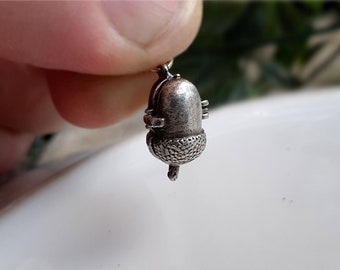 May include: A small silver-toned metal acorn pendant featuring a smooth, rounded top section and a textured, cross-hatched cap base. The charm includes a small metal loop at the top for attachment and exhibits a weathered, metallic finish throughout.