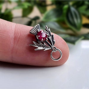 May include: Silver charm featuring a red ruby set in a stylized thistle design. The charm has a loop for attaching to a bracelet or necklace.