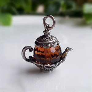 Może przedstawiać: Srebrny charm w kształcie czajnika z brązowym, fasetowanym szklanym korpusem. Charm ma skomplikowane detale i pętlę do przymocowania do bransoletki lub naszyjnika.