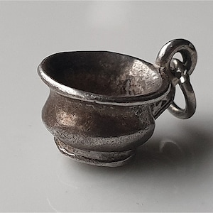May include: A silver charm shaped like a small, round, silver pot with a handle. The pot has a textured surface and a slightly rounded bottom.