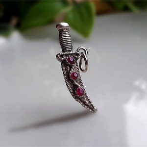 Könnte beinhalten: Ein silberner Dolch-Anhänger mit strukturiertem Griff und drei roten Edelsteinen. Die Klinge hat einen dekorativen Rand. Der Anhänger hat eine kleine Schlaufe zum Befestigen an einer Halskette oder einem Armband.