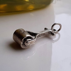 May include: A small, silver-toned charm in the shape of a paint roller. The roller is cylindrical with a handle and a loop for attaching to a bracelet or necklace. The charm is likely made of metal and has a vintage or antique appearance.