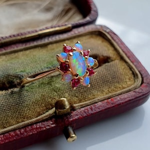 Könnte beinhalten: Ein goldener Ring mit einem ovalen Opal-Mittelstein, umgeben von roten Edelsteinen und kleineren Opalen. Der Ring ist in einem goldenen Band gefasst und wird in einer Vintage-Schmuckschatulle mit roter Außenseite und grünem Innenraum präsentiert.
