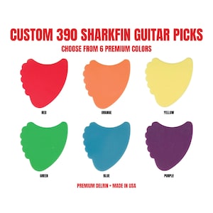 May include: Custom 390 Sharkfin guitar picks in six colours: red, orange, yellow, green, blue, and purple. The text reads: CUSTOM 390 SHARKFIN GUITAR PICKS, CHOOSE FROM 6 PREMIUM COLOURS, RED, ORANGE, YELLOW, GREEN, BLUE, PURPLE, PREMIUM DELRIN • MADE IN USA.