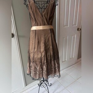Puede incluir: Vestido marr&oacute;n sin mangas con cuello en V y faj&iacute;n beige. El vestido tiene un patr&oacute;n decorativo en el dobladillo y est&aacute; adornado con peque&ntilde;os detalles dispersos. El vestido se exhibe en un maniqu&iacute; de metal negro.