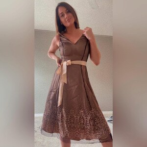 Puede incluir: Un vestido de fiesta marr&oacute;n con cuello en V y tirantes finos. El vestido presenta un cintur&oacute;n de lazo beige y un estampado decorativo en el dobladillo. Tiene una capa transparente y una falda a la rodilla.