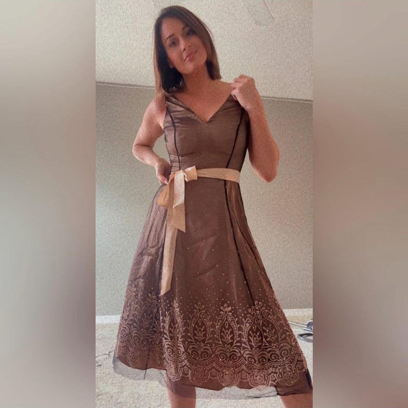 Puede incluir: Un vestido de fiesta marr&oacute;n con cuello en V y tirantes finos. El vestido presenta un cintur&oacute;n de lazo beige y un estampado decorativo en el dobladillo. Tiene una capa transparente y una falda a la rodilla.