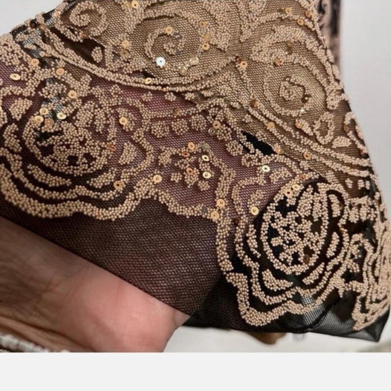 Puede incluir: Primer plano de una secci&oacute;n de tela con intrincados patrones florales. La tela presenta una base de malla negra superpuesta con un dise&ntilde;o detallado en cuentas y lentejuelas doradas y bronceadas. El dise&ntilde;o incluye motivos florales arremolinados.