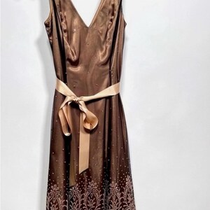 Puede incluir: Vestido marr&oacute;n sin mangas con escote en V y cinta marr&oacute;n claro atada a la cintura. El vestido tiene un dobladillo estampado y peque&ntilde;os adornos dispersos. La tela tiene un ligero brillo y el vestido parece estar hecho de un material ligero.