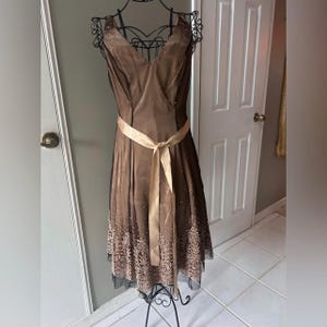 Puede incluir: Un vestido de c&oacute;ctel marr&oacute;n con cuello en V y cintur&oacute;n de cinta de sat&eacute;n. El vestido presenta un dobladillo decorativo y se exhibe en un maniqu&iacute; de metal negro. El vestido tiene una superposici&oacute;n transparente.