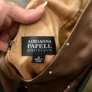 Puede incluir: Primer plano de una etiqueta de vestido de Adrianna Papell Boutique. La etiqueta es negra con texto blanco y dice "Adrianna Papell Boutique" y "Made in China". El vestido es talla 4. El vestido tiene una superposici&oacute;n de malla marr&oacute;n con peque&ntilde;os acentos florales blancos.