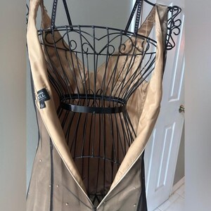 Puede incluir: Un vestido formal en color neutro, colgado de un maniqu&iacute; de alambre negro. El vestido tiene cuello en V y tirantes finos. El vestido est&aacute; hecho de una capa exterior negra transparente sobre un forro beige. Una etiqueta es visible.