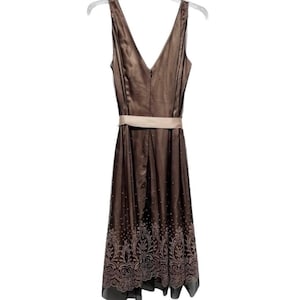 Puede incluir: Vestido marr&oacute;n sin mangas con cuello en V y cintur&oacute;n beige. El vestido tiene un dise&ntilde;o estampado en la parte inferior y est&aacute; colgado de una percha blanca.