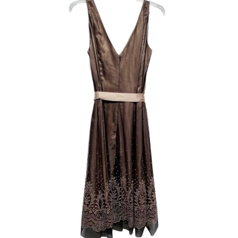 Puede incluir: Vestido marr&oacute;n sin mangas con cuello en V y cintur&oacute;n beige. El vestido tiene un dise&ntilde;o estampado en la parte inferior y est&aacute; colgado de una percha blanca.