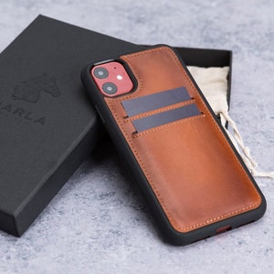 May include: A brown leather phone case with a black trim and two card slots. The case has a red camera cutout and the word "MARLA" is embossed on the back of the case.
