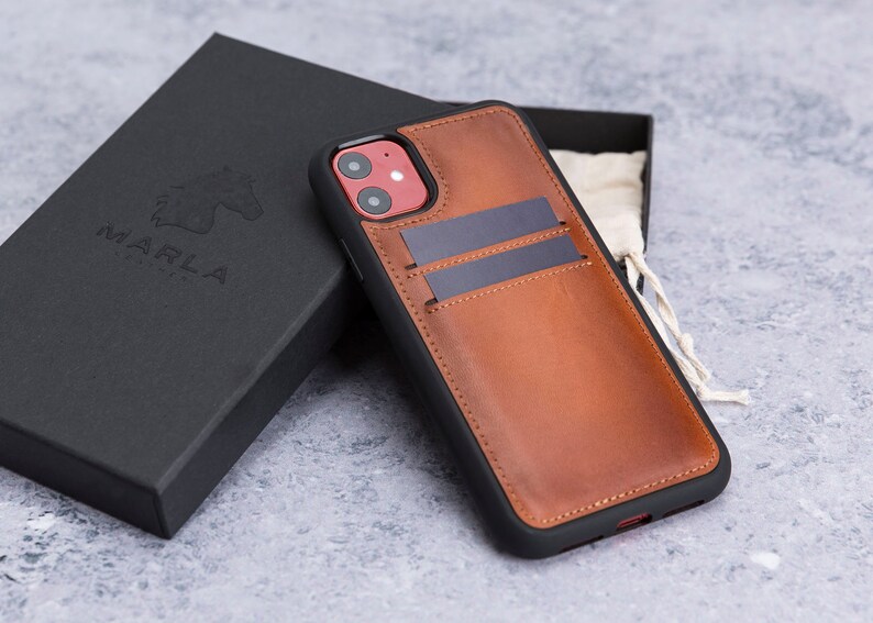 May include: A brown leather phone case with a black trim and two card slots. The case has a red camera cutout and the word "MARLA" is embossed on the back of the case.