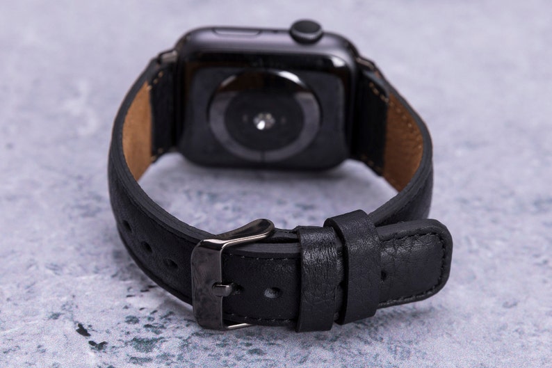 May include: Black leather watch band with a black metal buckle. The band is designed for an Apple Watch.