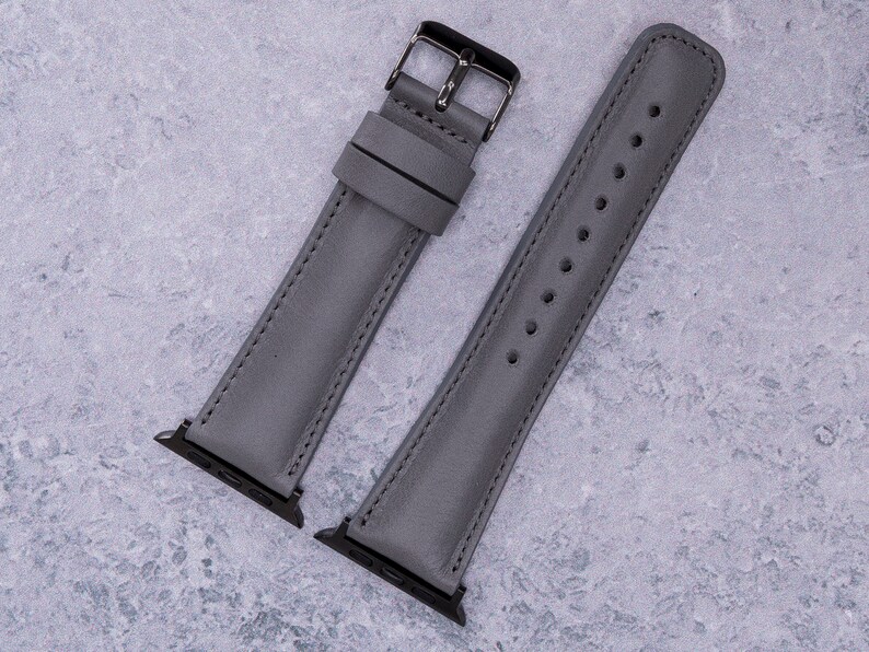 May include: Two gray leather watch bands with silver hardware. The bands are laid out flat on a gray textured surface.