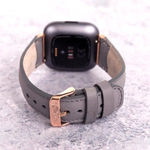 May include: A gray leather watch band with a rose gold buckle and a black smartwatch face.