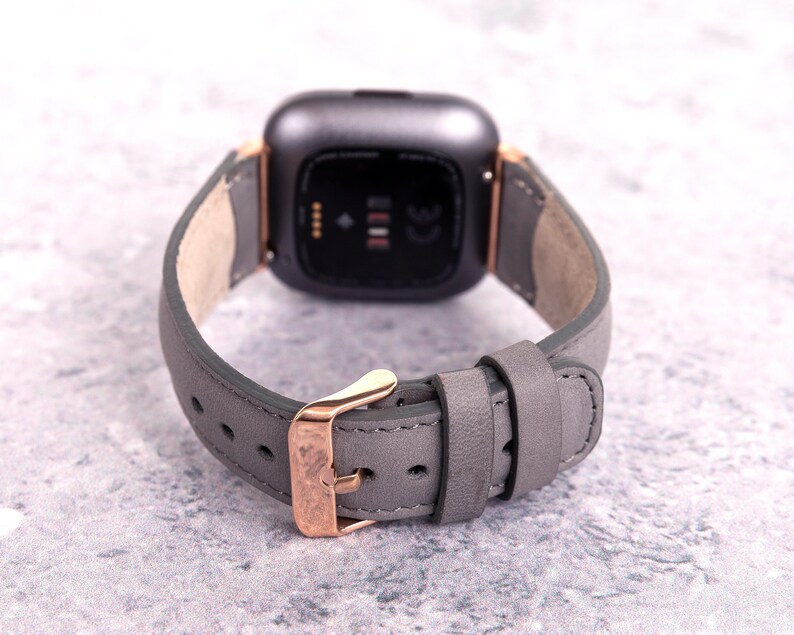 May include: A gray leather watch band with a rose gold buckle and a black smartwatch face.