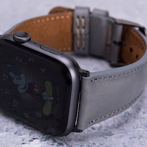 May include: A gray leather watch band with a black Apple Watch with a Mickey Mouse watch face. The band has a brown leather lining.