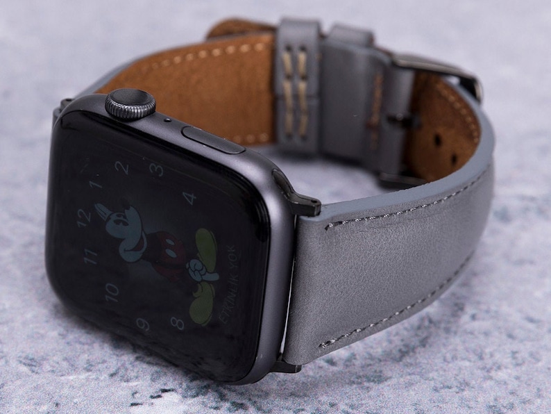 May include: A gray leather watch band with a black Apple Watch with a Mickey Mouse watch face. The band has a brown leather lining.