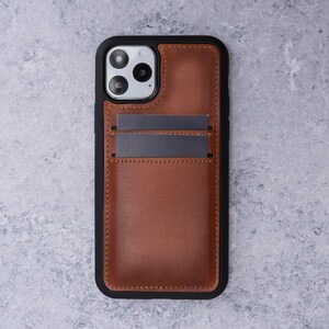 May include: A brown leather phone case with a black frame and a built-in card holder. The case has a camera cutout and a textured leather finish.