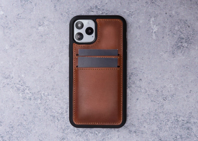May include: A brown leather phone case with a black frame and a built-in card holder. The case has a camera cutout and a textured leather finish.