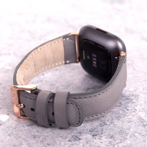 May include: A gray leather watch band with rose gold hardware. The band is designed for a smartwatch with a square face.