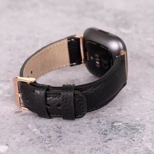 May include: Black leather watch band with a rose gold buckle and a gray smartwatch. The band has a textured leather finish.