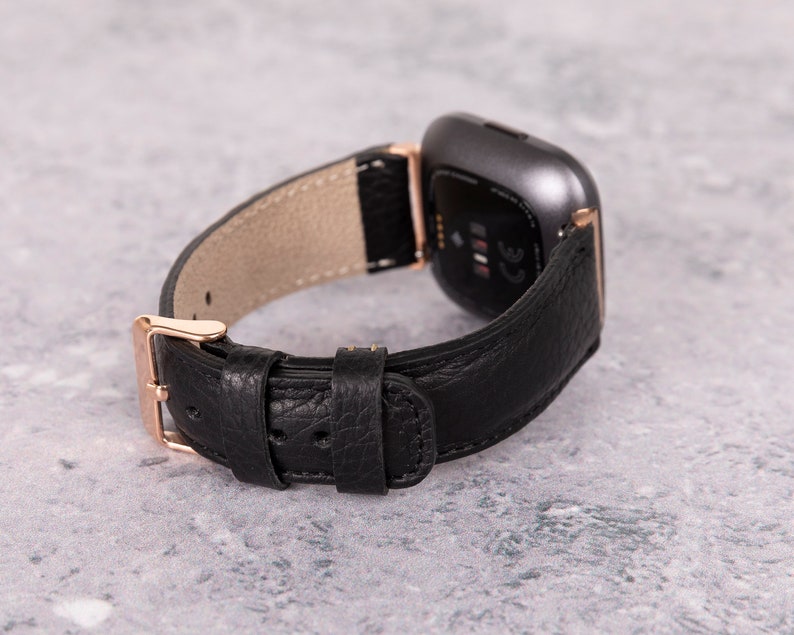 May include: Black leather watch band with a rose gold buckle and a gray smartwatch. The band has a textured leather finish.