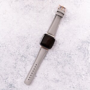 May include: A gray leather watch band with a black square watch face. The watch band has a rose gold buckle.