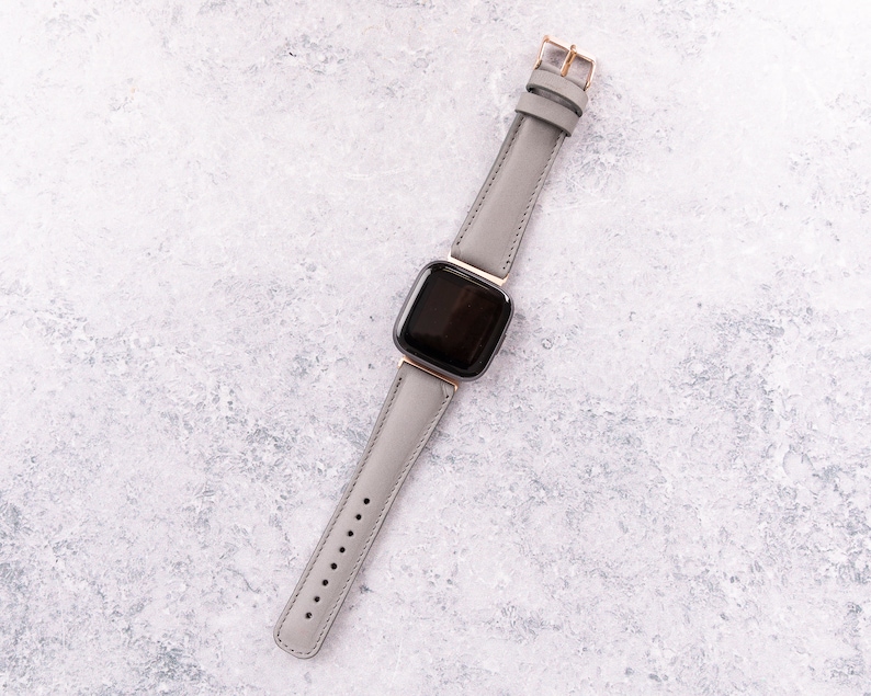 May include: A gray leather watch band with a black square watch face. The watch band has a rose gold buckle.