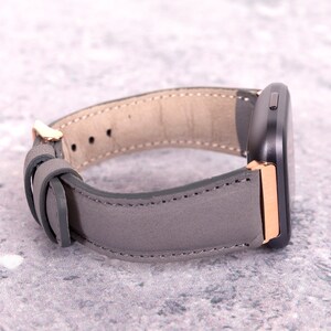 May include: A gray leather watch band with rose gold accents. The band is designed for a smartwatch with a square face.