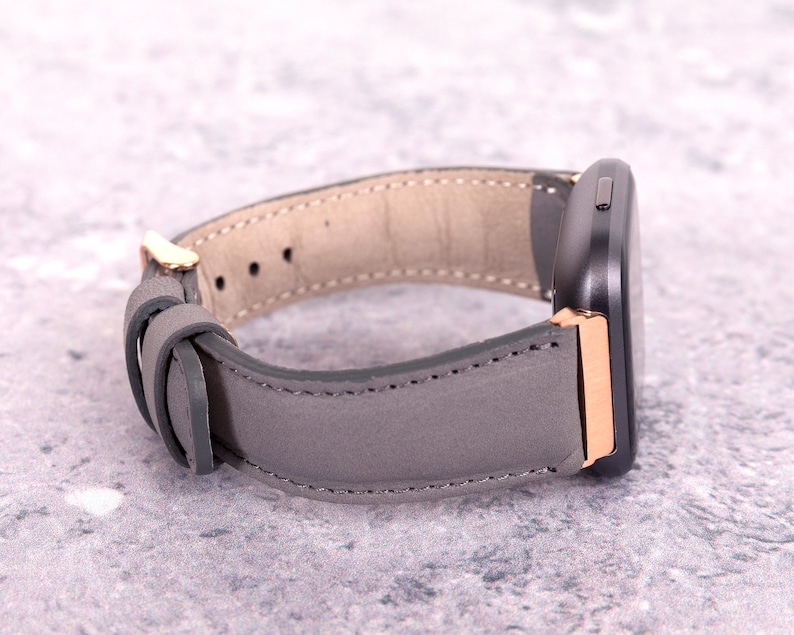 May include: A gray leather watch band with rose gold accents. The band is designed for a smartwatch with a square face.