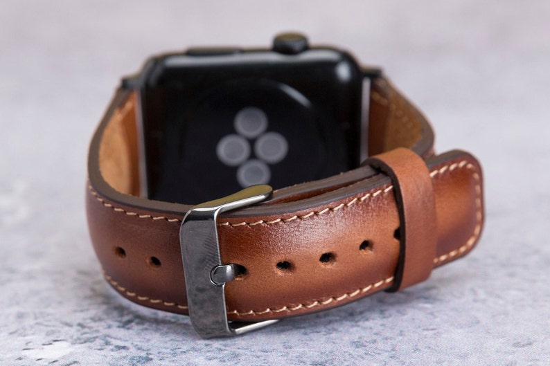May include: A brown leather watch band with a silver buckle and stitching. The band is attached to a black smartwatch with a black screen.