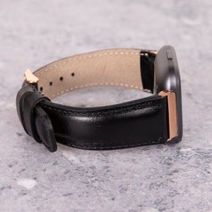 May include: A black leather watch band with a rose gold buckle and a dark gray watch face.
