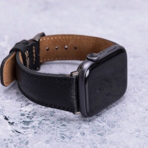 May include: A black leather watch band with a brown leather lining. The band is attached to a black Apple Watch.