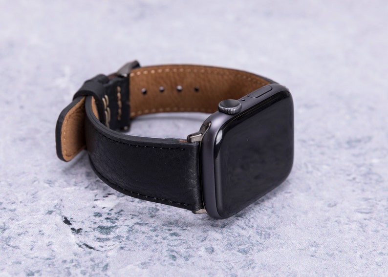 May include: A black leather watch band with a brown leather lining. The band is attached to a black Apple Watch.