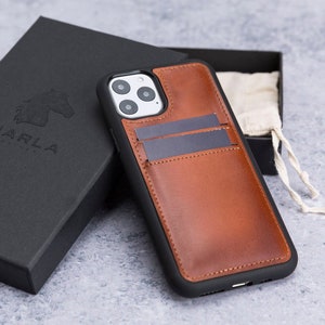 May include: A brown leather phone case with a built-in card holder. The case has a black trim and a cutout for the camera lens. The case is sitting on a gray surface.