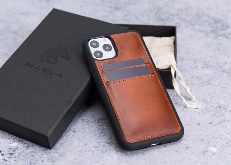May include: A brown leather phone case with a built-in card holder. The case has a black trim and a cutout for the camera lens. The case is sitting on a gray surface.