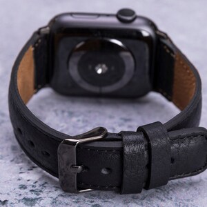 以下が含まれることがあります： 銀色のバックルが付いた黒い革製の腕時計バンド。バンドはApple Watch用に設計されています。