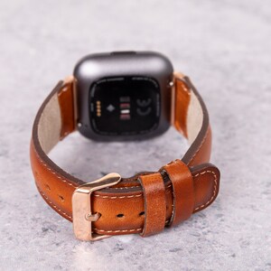 Puede incluir: Una correa de reloj de cuero marr&oacute;n con una hebilla dorada y una esfera de reloj inteligente plateada.